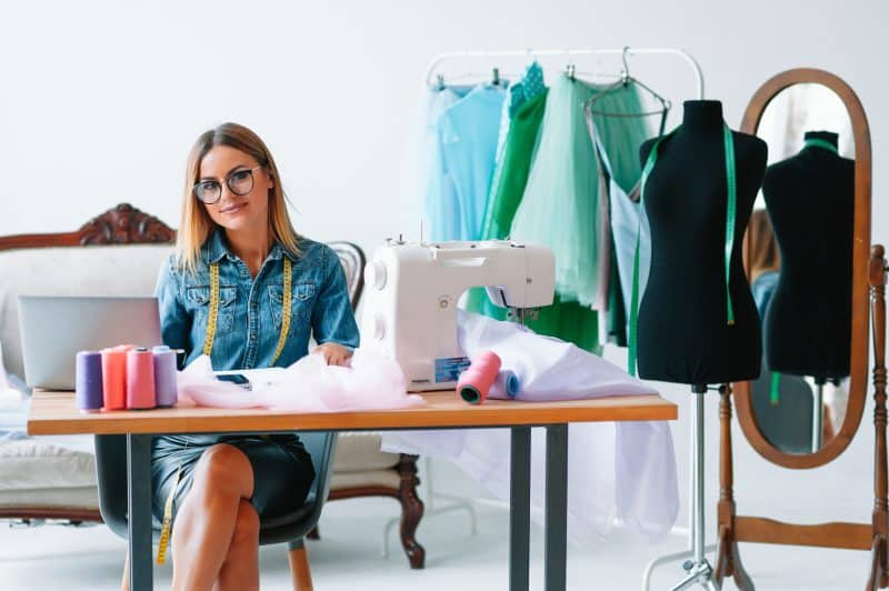 Smiling fashion designer looking at camera at workplace, dressmaker, needlewoman or tailor shop owner sitting at desk with color swatches pantone and embroidery design sketches on the wall, portrait
